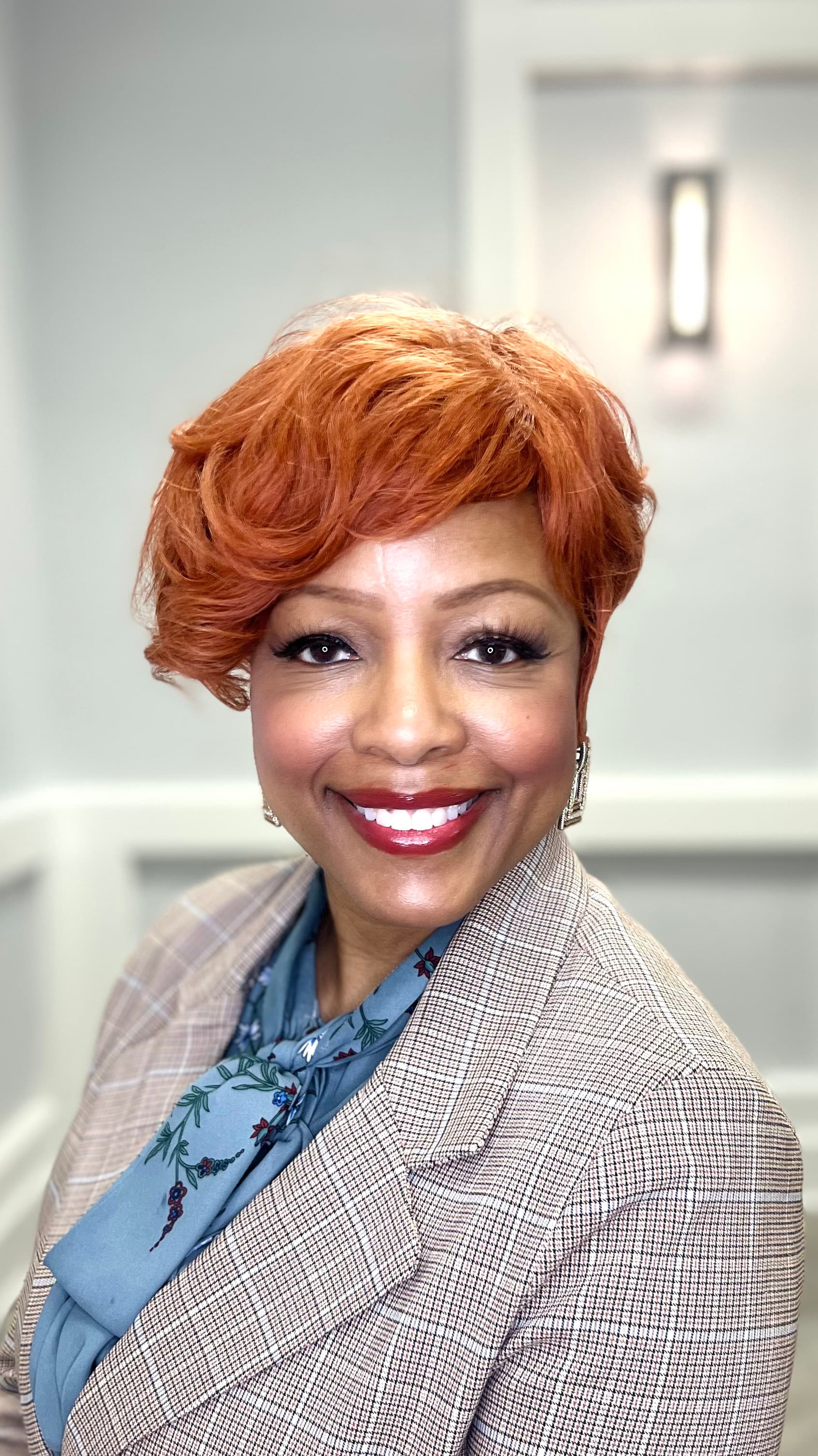 Smiling woman with short reddish hair wearing a checkered blazer and blue floral blouse.